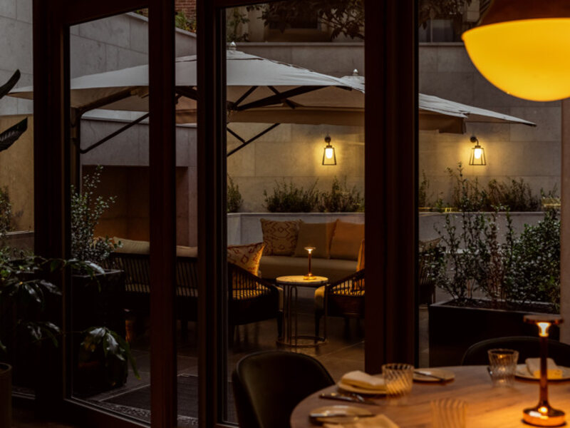 A laid table at Le Petit Bon Bon in Brussels in the evening, looking through glass doors onto a lit courtyard terrace with parasols and wall lanterns