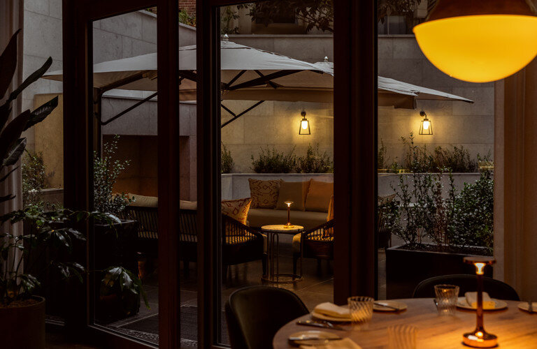 A laid table at Le Petit Bon Bon in Brussels in the evening, looking through glass doors onto a lit courtyard terrace with parasols and wall lanterns