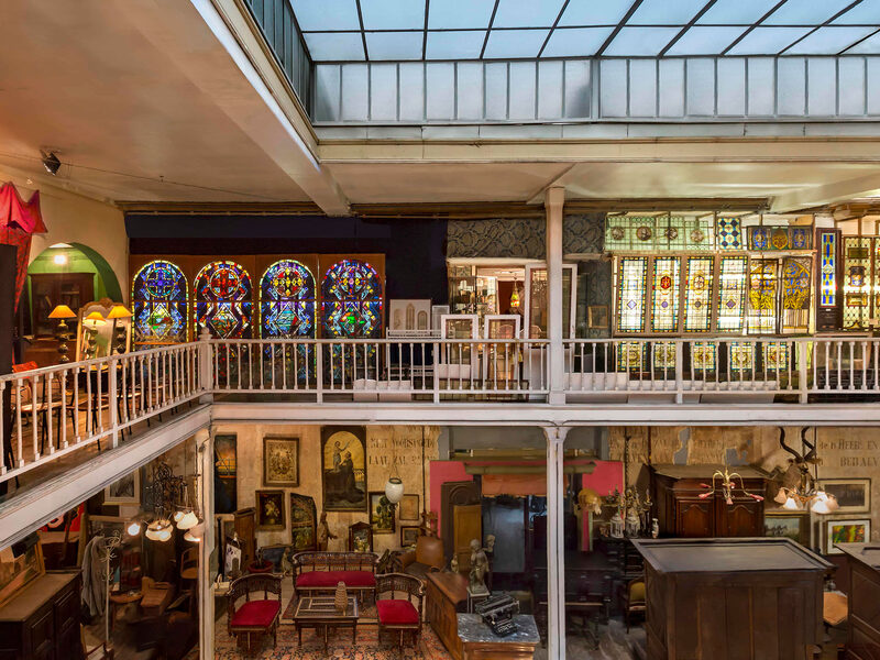 The interior of Passage 125 in Brussels seen from an upper gallery, panels of stained glass ranged along the walls under a glazed roof, with chandeliers, framed paintings and dark wooden furniture on the floor below