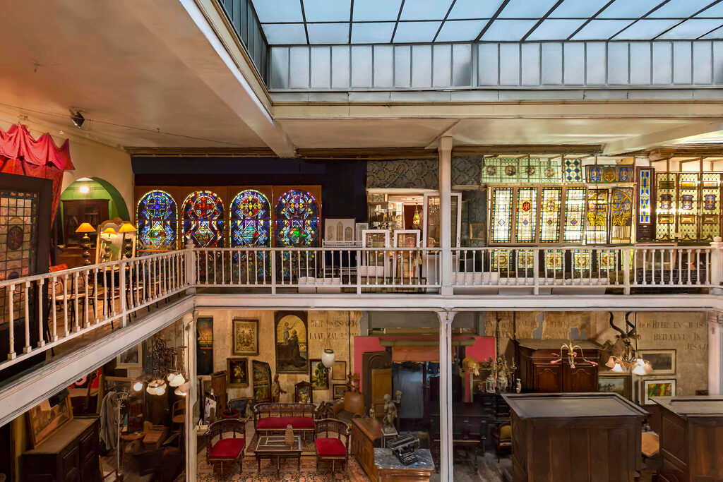 The interior of Passage 125 in Brussels seen from an upper gallery, panels of stained glass ranged along the walls under a glazed roof, with chandeliers, framed paintings and dark wooden furniture on the floor below