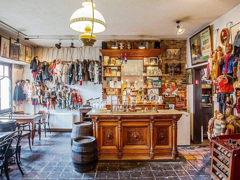 The interior of Estaminet Toone in Brussels, marionettes in costume hanging along the walls and above a carved oak bar, with a brick floor, bentwood chairs and a wooden beer barrel