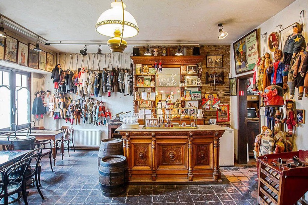 The interior of Estaminet Toone in Brussels, marionettes in costume hanging along the walls and above a carved oak bar, with a brick floor, bentwood chairs and a wooden beer barrel