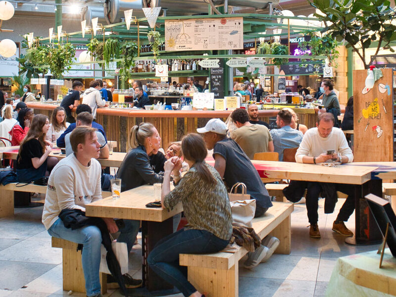 The main hall at Wolf in Brussels, people eating at long wooden communal tables around a central bar, with spherical lamps hanging under a glazed roof