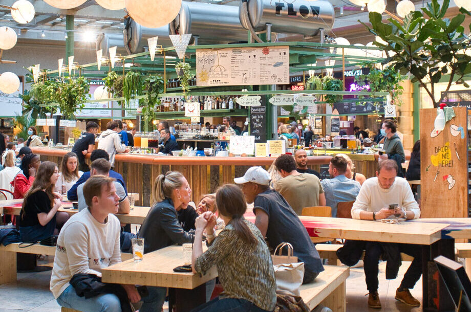 The main hall at Wolf in Brussels, people eating at long wooden communal tables around a central bar, with spherical lamps hanging under a glazed roof