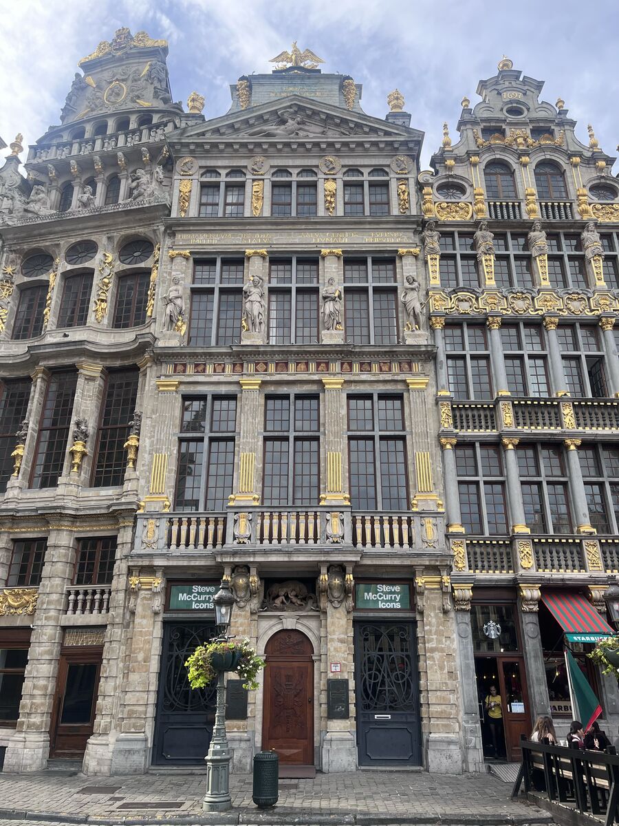 La Louve on the north-west row of the Grand-Place, statues standing in niches between its upper windows beneath a triangular pediment, with Steve McCurry Icons signs either side of the door at street level and Le Sac's café beyond to the right.