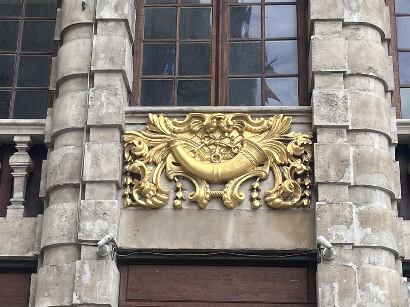 The gilded emblem of Le Cornet: a curved horn wrapped in acanthus scrollwork, set above the door between rusticated stone columns.