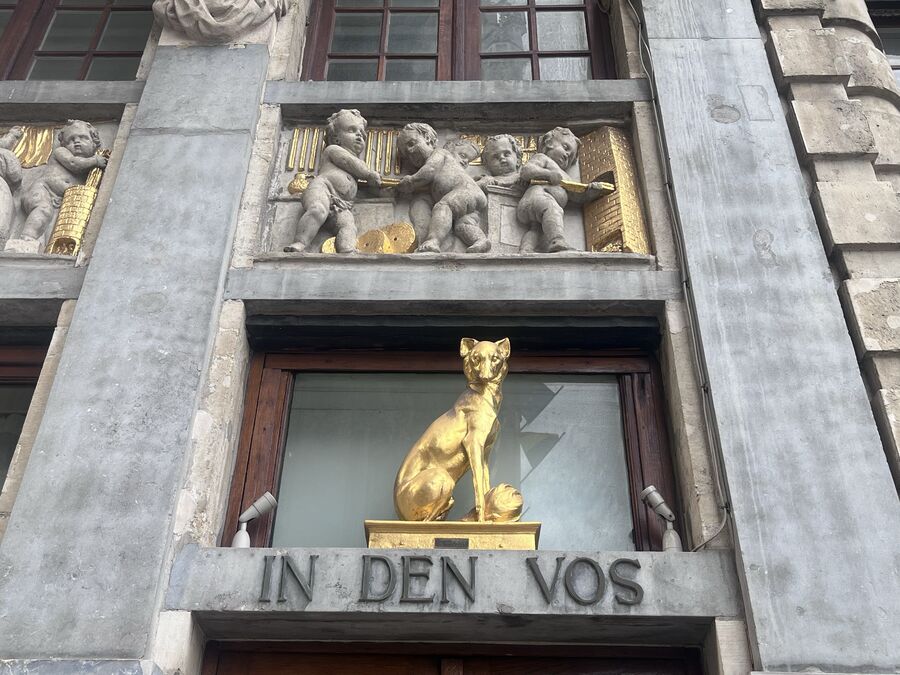 A gilded fox seated above the doorway of Le Renard, with IN DEN VOS carved into the lintel beneath it and putti holding a golden disc above.
