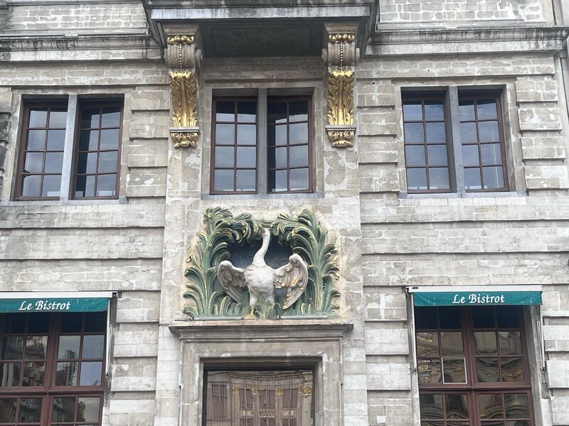 Le Cygne on the Grand-Place, the swan carved above its door in a spray of green foliage and the date Anno 1698 on the gable above, with Le Bistrot's windows either side and visitors sitting on the steps.