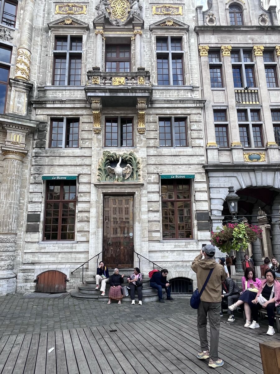 Le Cygne on the Grand-Place, the swan carved above its door in a spray of green foliage and the date Anno 1698 on the gable above, with Le Bistrot's windows either side and visitors sitting on the steps.