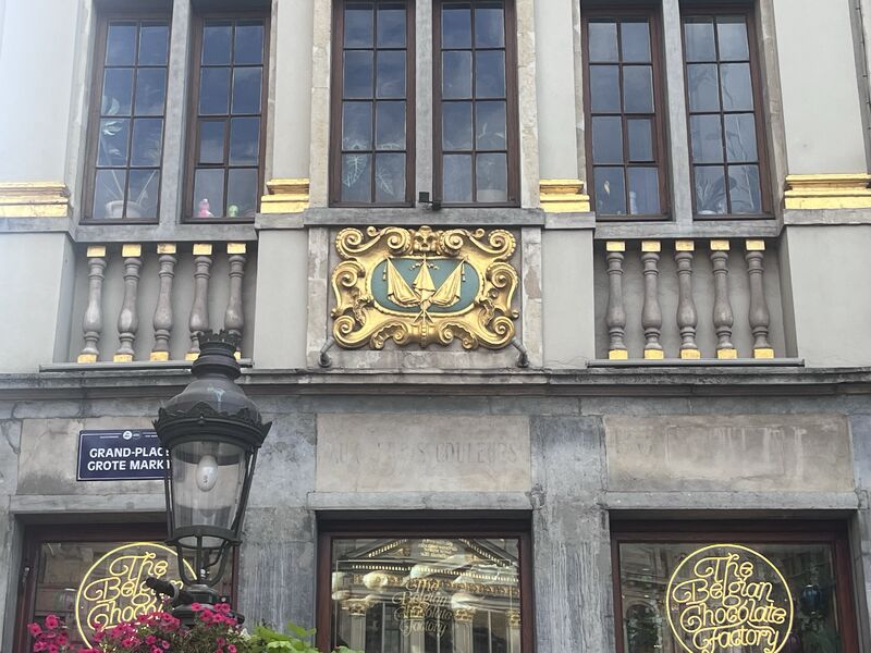 A gilded emblem in a scrolled cartouche between stone balusters on the facade of Le Mont Thabor, above the chocolate shop at street level.