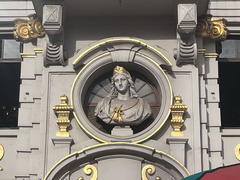 The bust of Saint Barbara set in the circular transom window above the entrance of La Chaloupe d'Or, ringed in gold and flanked by gilded urns on the grey stone.