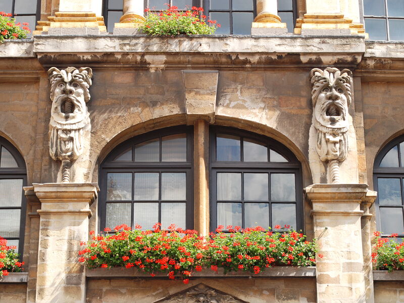 Two carved grotesque masks on corbels at mezzanine level on the Maison du Pigeon, either side of an arched window, with red geraniums in the window boxes below.